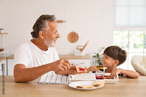 Little boy and his grandfather making sandwiches with jam at table in kitchen