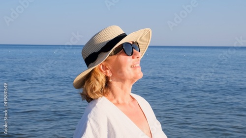 Portrait of a contented 50 year old woman wearing a straw hat and sunglasses enjoying the sun against the blue sea. Summer, vacation, vacation, active retirees