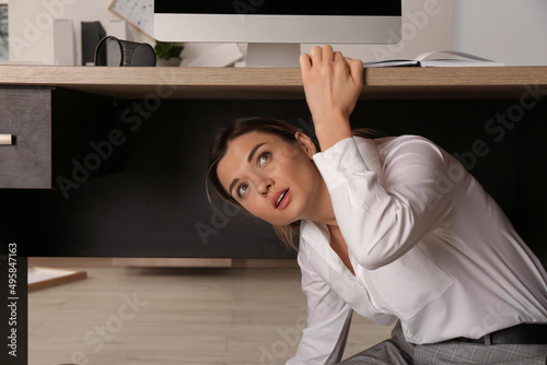 Quadro em tela Scared young woman hiding under office desk during earthquake