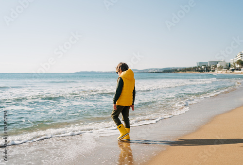 Boy in yellow rubber boots playing with water and sand at the beach. School kid touching water at autumn winter sea. Child having fun with waves at the shore. Spring Holiday vacation concept.