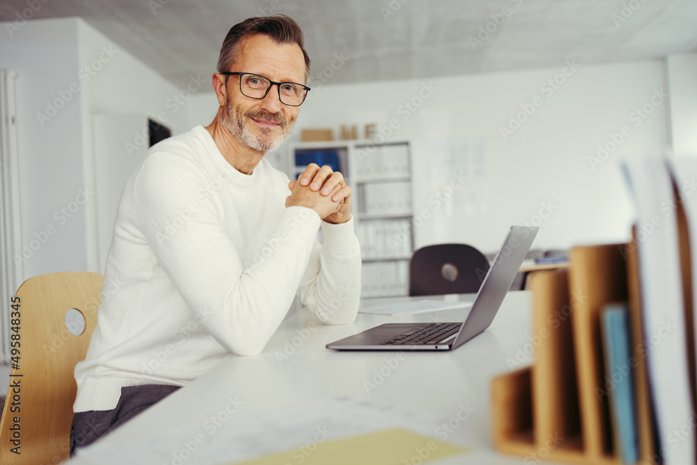 casual smiling man sitting at his desk in front of a laptop in his office casual smiling man sitting at his desk in front of a laptop in his office