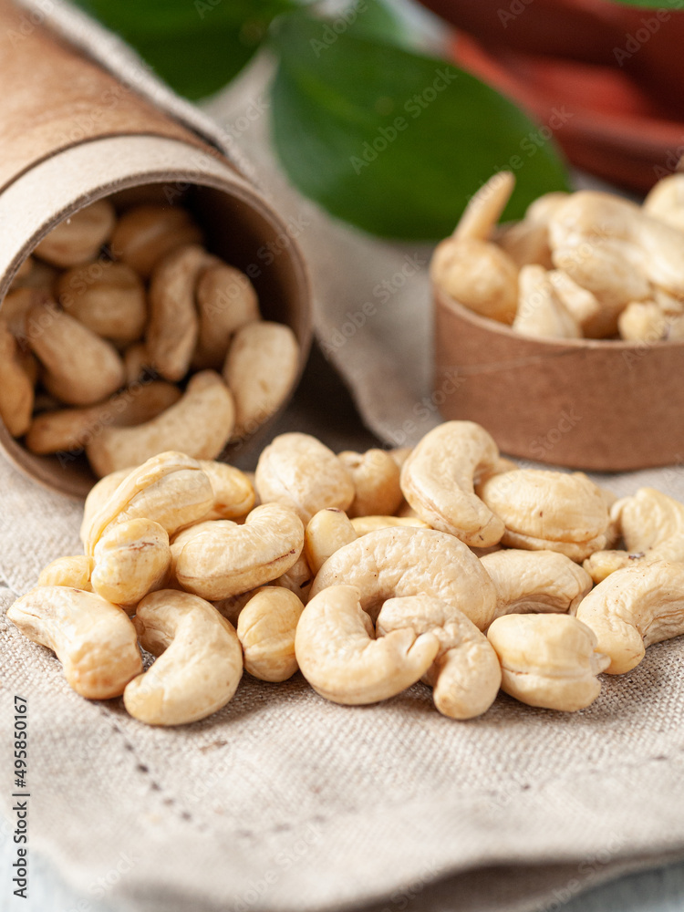 Peeled cashews are scattered on the table. There is a cardboard box next to it. Gray linen background. Close-up. Peeled cashews are scattered on the table. There is a cardboard box next to it. Gray linen background. Close-up.
