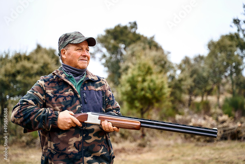 Male hunter in camouflage outerwear and cap carrying a gun while hanging out in the autumn field