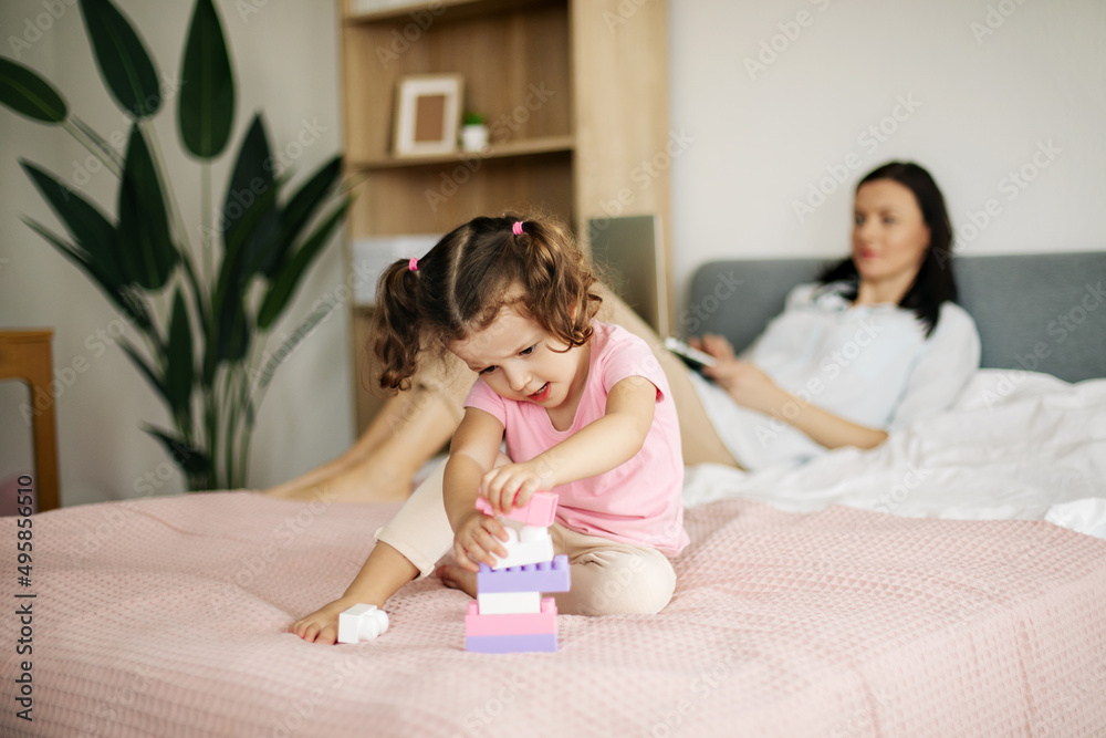 A cute girl is playing with a constructor on the bed while her mom is working on a laptop A cute girl is playing with a constructor on the bed while her mom is working on a laptop
