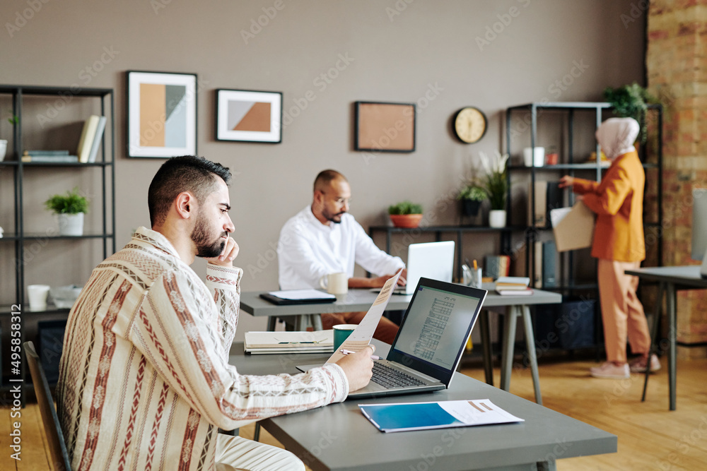 Obraz premium Serious young male economist analyzing financial papers in front of laptop by workplace in office against two busy coworkers
