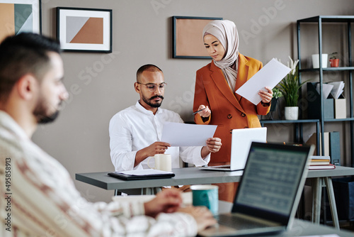 Young Muslim female and male economists discussing financial papers while businesswoman pointing at one held by colleague