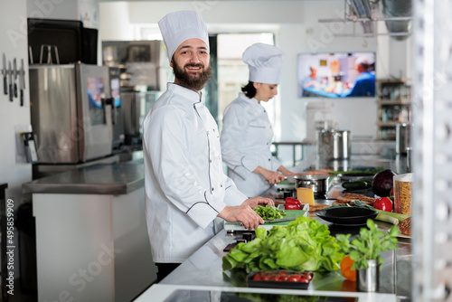 Fototapeta Naklejka Na Ścianę i Meble -  Portrait of positive smiling head chef standing in professional restaurant kitchen while looking at camera. Confident man wearing food industry white uniform while preparing delicious meal.
