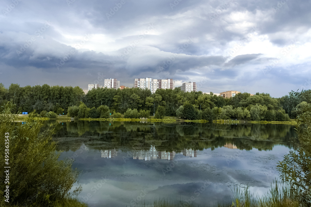 Summer city landscape with calm lake water surface with cloudy sky and multi- storey residential buildings reflected on water Summer city landscape with calm lake water surface with cloudy sky and multi- storey residential buildings reflected on water