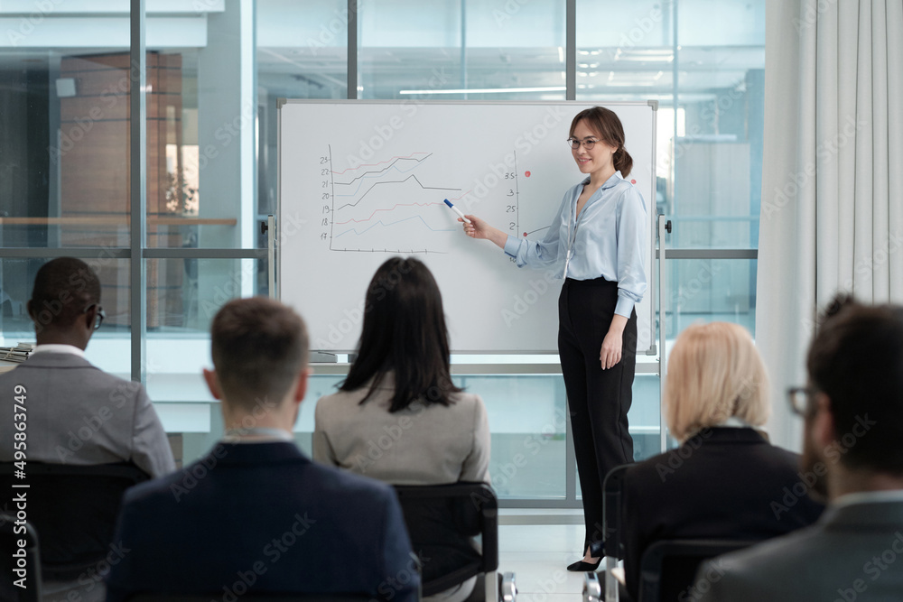 Young successful female broker pointing at financial graphs on whiteboard while standing in front of audience in lecture hall