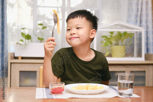 Cute happy smiling Asian 5 yers old kindergarten boy child using fork eating French fries potato chips with ketchup, sitting at the table at home or restaurant, Unhealthy Kids Foods concept