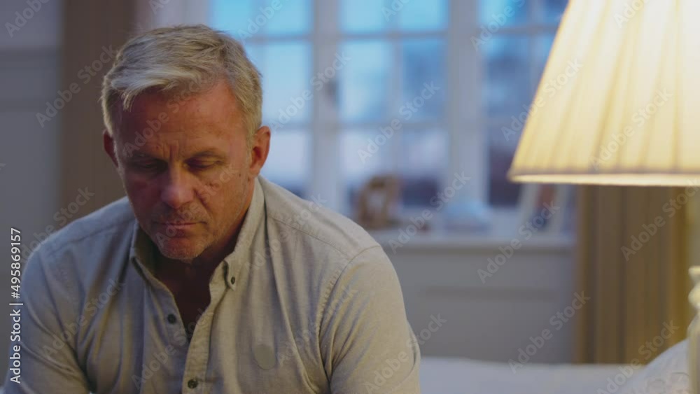Depressed mature man at home sitting in bedroom in evening looking sad- shot in slow motion Depressed mature man at home sitting in bedroom in evening looking sad- shot in slow motion