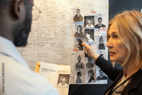 Blond confident female detective pointing at photo of suspect on board over map while matching facts of serial crimes with colleague photo