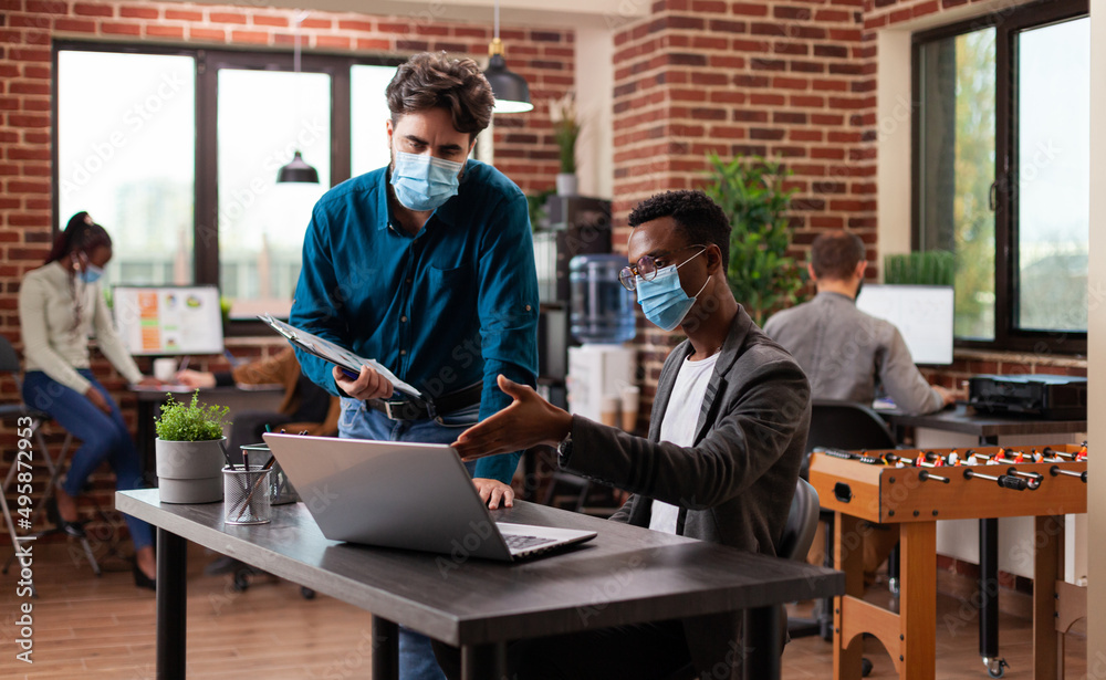 Diverse businessmen working at marketing project analyzing company graph discussing business ideas in brick wall startup office. Multi-ethnic team wearing medical face mask against covid19 Diverse businessmen working at marketing project analyzing company graph discussing business ideas in brick wall startup office. Multi-ethnic team wearing medical face mask against covid19