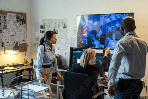 Head of safeguard department and her subordinates looking at security camera while standing in front of large screen in office