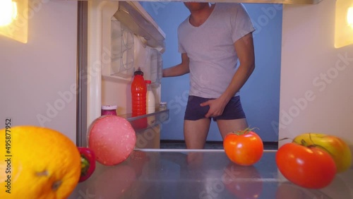 young man in underpants without pants opens refrigerator at night, looks inside, scratches his penis and egg, yawns, covers his mouth, takes a tomato. concept of night meal and snack. Night food. photo