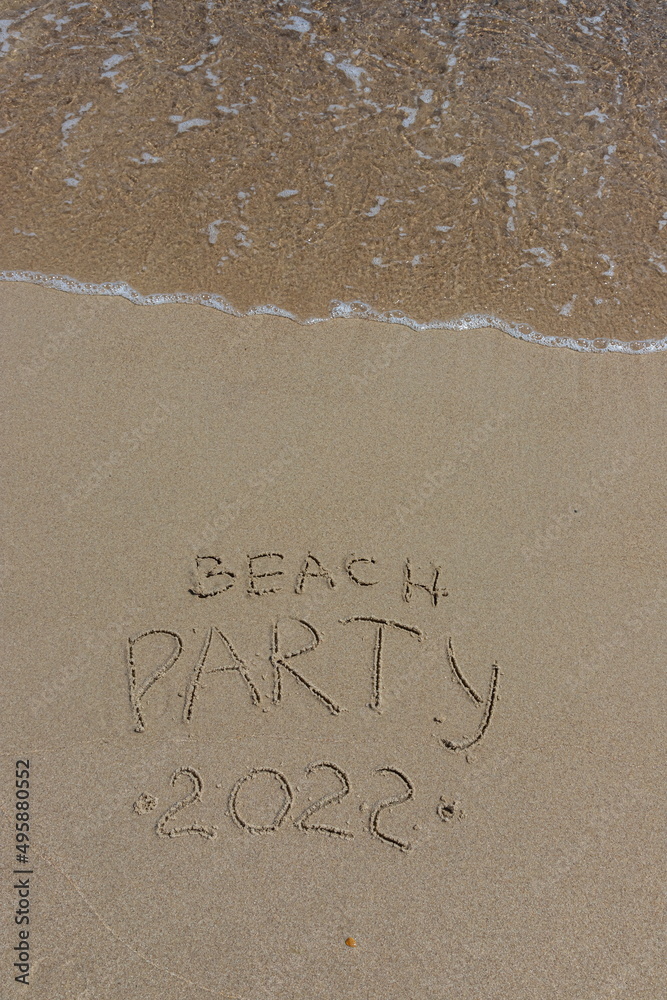Fototapeta premium The inscription beach party 2022 on the sand by the water and the rising wave, seashore beach vacation by the sea