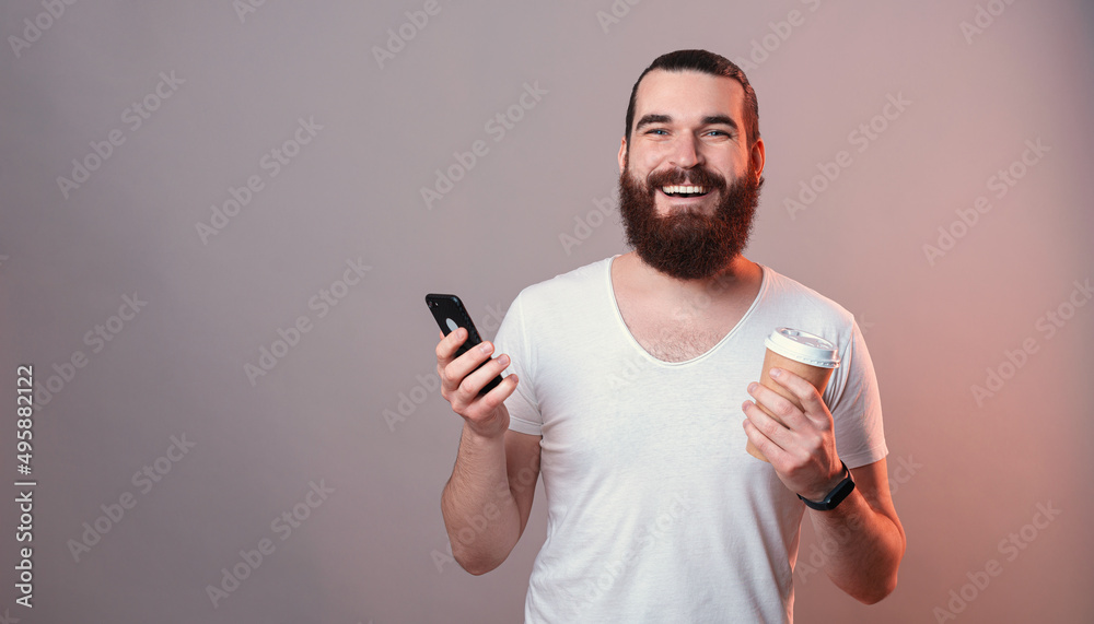 Smiling young man with beard holding cup of coffee to go and using smarpthone. Smiling young man with beard holding cup of coffee to go and using smarpthone.