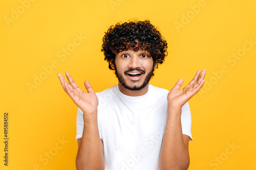 Surprised confused indian or arabian guy with curly hair, wearing white t-shirt, looking at camera in disbelief, spreading his arms to the sides, shrugging, standing over isolated orange background