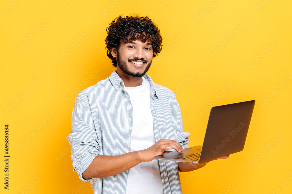 Attractive contented indian or arabian curly haired guy dressed in casual wear, holding an open laptop in his hand, looks friendly smiling at the camera, while standing on isolated orange background Attractive contented indian or arabian curly haired guy dressed in casual wear, holding an open laptop in his hand, looks friendly smiling at the camera, while standing on isolated orange background