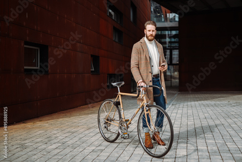 Young man with bicycle holding a smartphone front of building looking to camera.