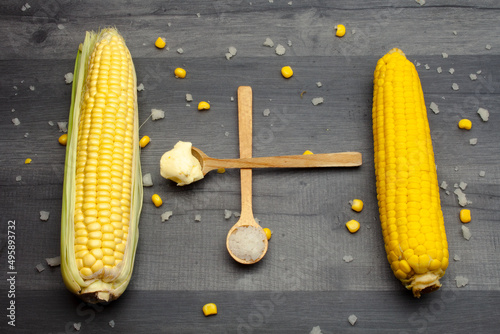 Two cobs of fresh and cooked sweet corn lying on a gray table surrounded by salt and butter on a wooden spoon.
