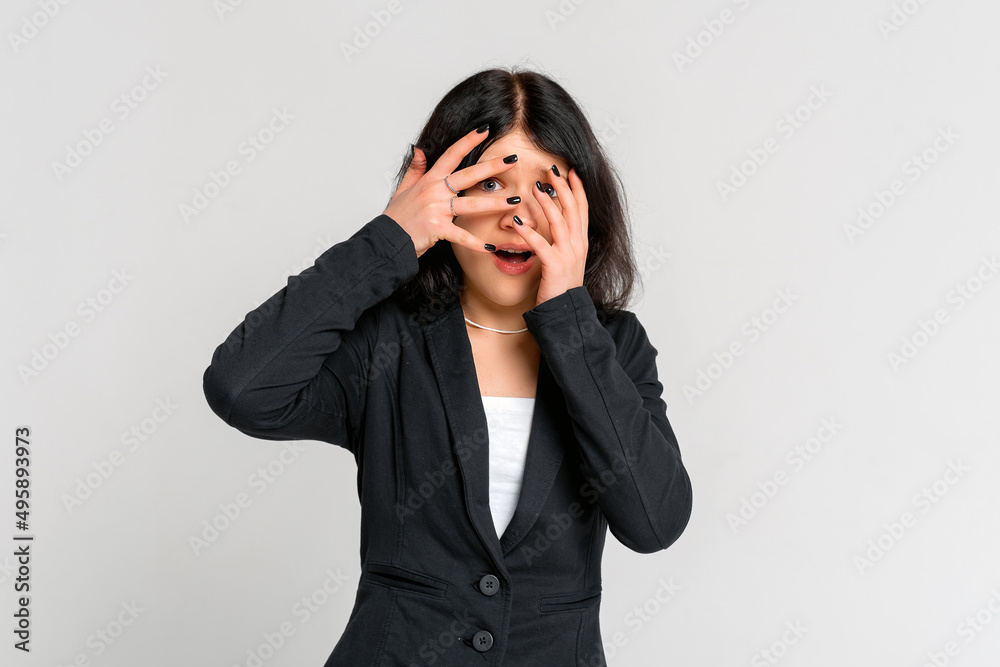 Scared teen girl hiding face in palms hands and peering out with one eye between her fingers, trembling fear, very terrified, standing in black blazer over grey background Scared teen girl hiding face in palms hands and peering out with one eye between her fingers, trembling fear, very terrified, standing in black blazer over grey background