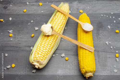 Two cobs of fresh and cooked sweet corn lying on a gray table surrounded by salt and butter on a wooden spoon.
