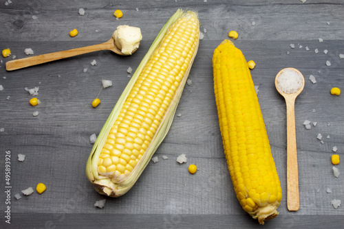 Two cobs of fresh and cooked sweet corn lying on a gray table surrounded by salt and butter on a wooden spoon.
