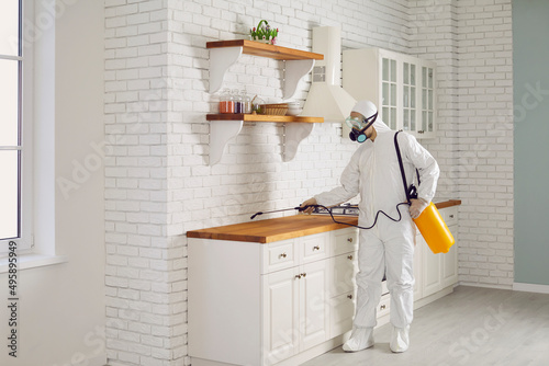 Exterminator fighting termites or cockroaches in the house. Pest control guy in mask and protective white suit spraying insecticide from sprayer bottle on cupboards and countertop in kitchen interior photo