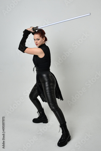 Full length portrait of pretty red haired female model wearing black futuristic scifi leather costume, holding a lightsaber sword weapon. Dynamic standing poses on a white studio background.