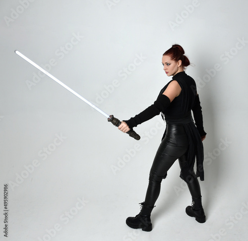 Full length portrait of pretty red haired female model wearing black futuristic scifi leather costume, holding a lightsaber sword weapon. Dynamic standing poses on a white studio background.