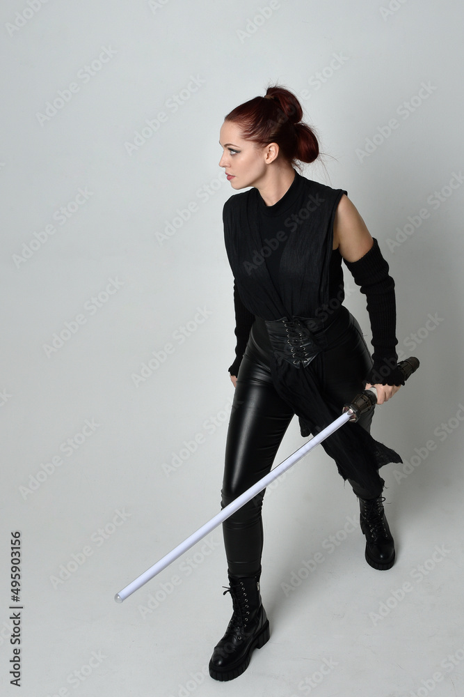 Full length portrait of pretty red haired female model wearing black futuristic scifi leather costume, holding a lightsaber sword weapon. Dynamic standing poses  on a white studio background.