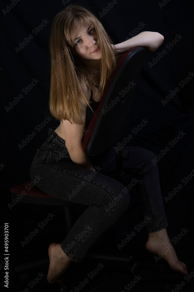 Sexy and beautiful young blonde with a slender body, long seductive legs posing in jeans on a chair, dark wall on the background Sexy and beautiful young blonde with a slender body, long seductive legs posing in jeans on a chair, dark wall on the background
