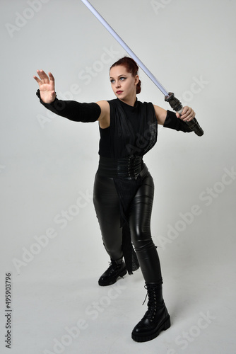 Full length portrait of pretty red haired female model wearing black futuristic scifi leather costume, holding a lightsaber sword weapon. Dynamic standing poses on a white studio background.