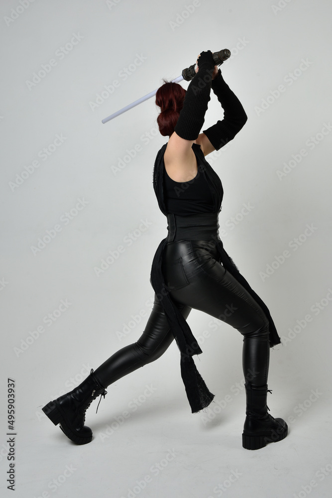 Full length portrait of pretty red haired female model wearing black futuristic scifi leather costume, holding a lightsaber sword weapon. Dynamic standing poses  on a white studio background.