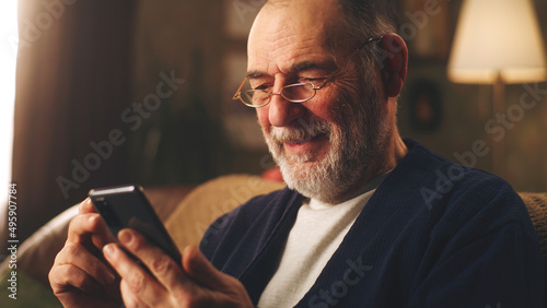 Elderly man smiling and watching funny videos on social networks using smartphone and resting in the evening sitting on the couch
