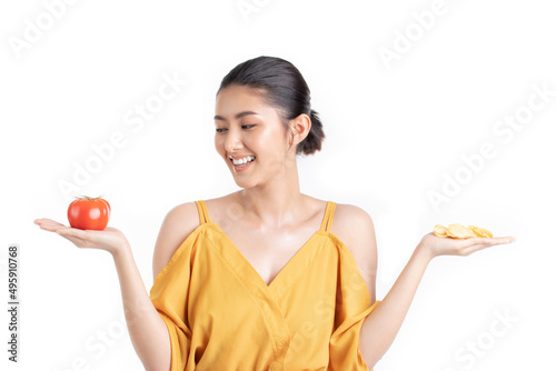 Attractive beautiful young asian woman in a pretty yellow dress choosing between fresh tomato and potato chips isolated on white background. Healthy diet food concept.