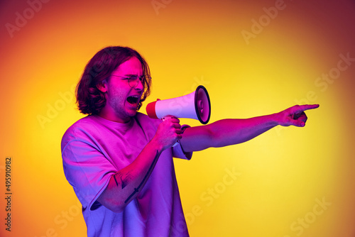 One young handsome man in white t-shirt shouting at megaphone isolated on orange background in neon light. Concept of emotions, fashion, youth