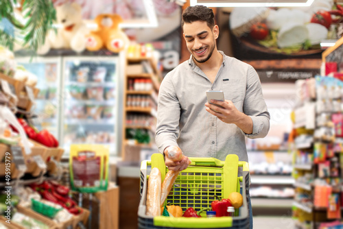 Middle Eastern Male Doing Grocery Shopping Using Cellphone In Supermarket