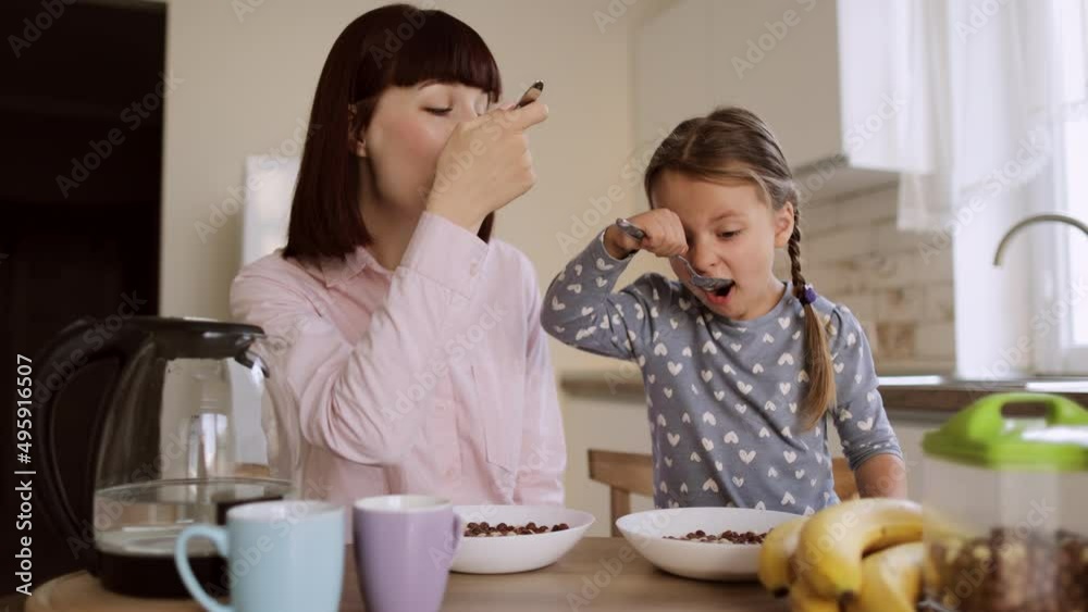 Young beautiful caucasian mother and her cute little girl having breakfast with chocolate corn balls with milk or yogurt sitting in a bright kitchen during the weekend. Young beautiful caucasian mother and her cute little girl having breakfast with chocolate corn balls with milk or yogurt sitting in a bright kitchen during the weekend.
