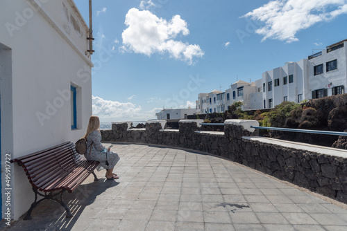 joven rubia sentada en Punta Mujeres, en las islas canarias de Lanzarote, sentada en un banco admirando la arquitectura con las casas blancas tradicionales en un día soleado con un cielo azul photo