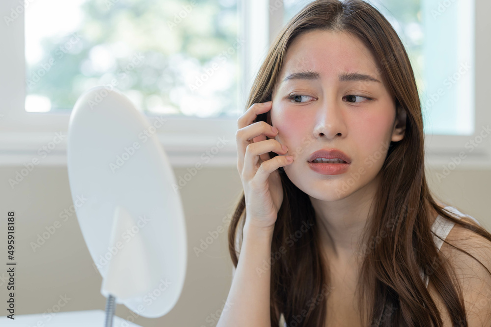 Dermatology, puberty asian young woman, girl looking into mirror, allergy presenting an allergic reaction from cosmetic, red spot or rash on face. Beauty care from skin problem by medical treatment. Dermatology, puberty asian young woman, girl looking into mirror, allergy presenting an allergic reaction from cosmetic, red spot or rash on face. Beauty care from skin problem by medical treatment.