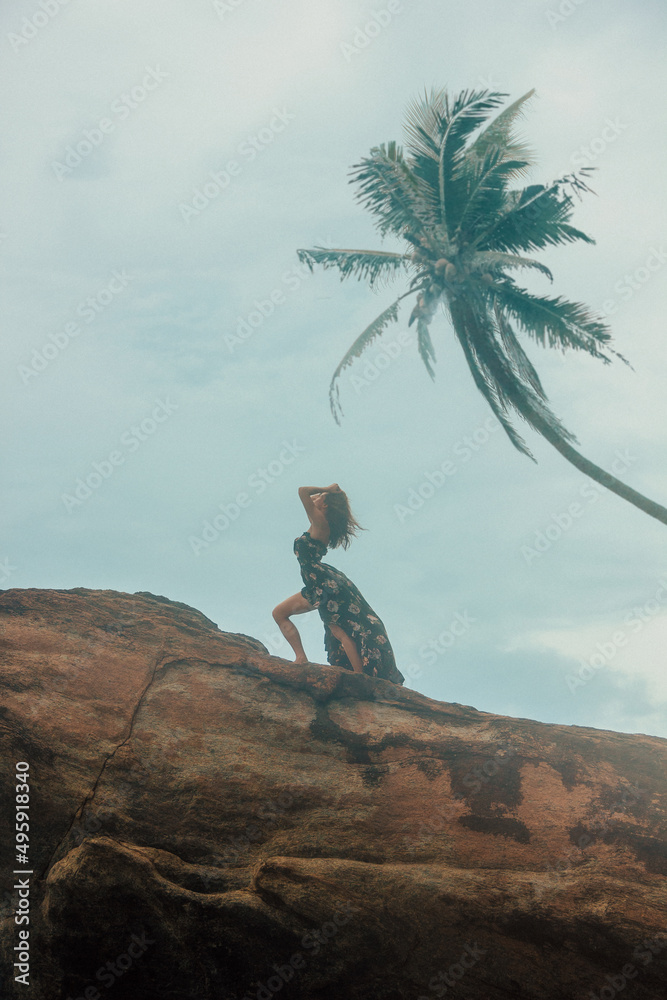 blogger leisure. young brunette girl in black stylish dress is standing fashion in profile with hands in hair on a huge rock on a blue sky background at sunset. travel lifestyle concept, free space