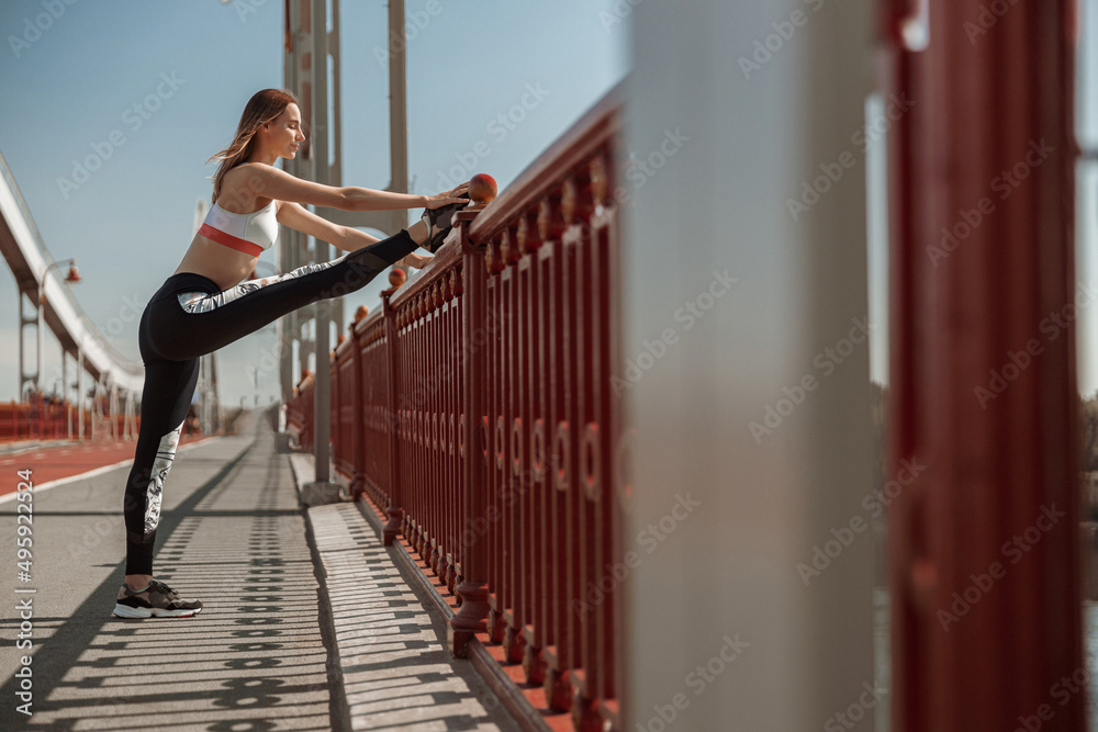Sportswoman in tracksuit stretches legs leaning onto railing on modern footbridge