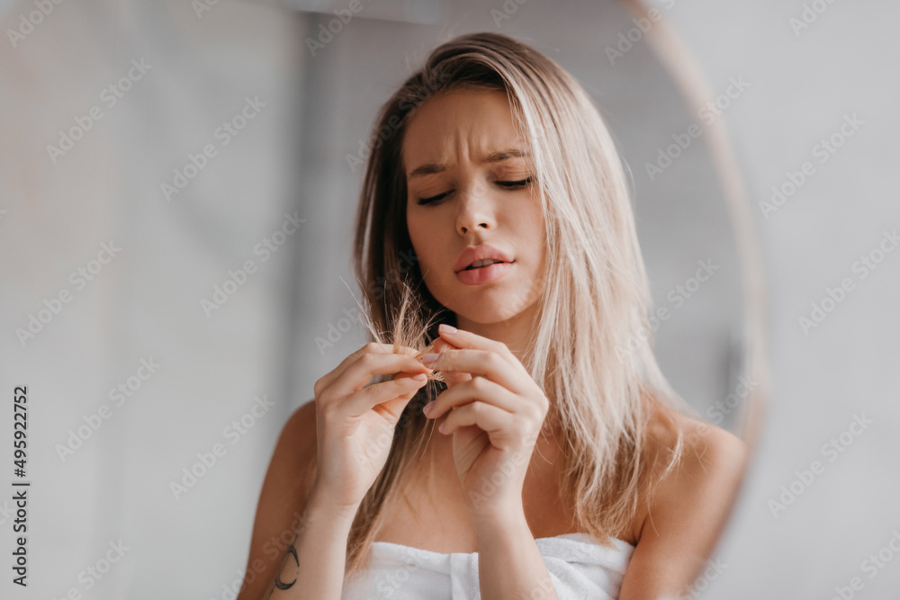 Beauty problems, dry split ends. Displeased woman in towel looking at ends of hair in morning, bathroom interior Beauty problems, dry split ends. Displeased woman in towel looking at ends of hair in morning, bathroom interior