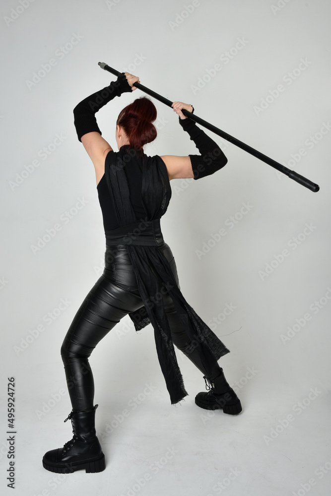 Full length portrait of pretty redhead female model wearing black futuristic scifi leather costume, holding a staff spear weapon. Dynamic standing pose facing away, backwards, white studio background. Full length portrait of pretty redhead female model wearing black futuristic scifi leather costume, holding a staff spear weapon. Dynamic standing pose facing away, backwards, white studio background.