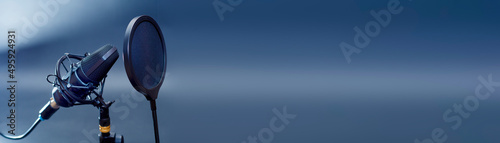 A narrow horizontal photo of a professional microphone with a pop filter in front of it. Desktop microphone for recording podcasts, songs and streaming against a dark background.