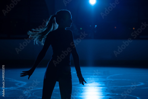 Young female figure skater is performing woman's single skating choreography on ice rink. Silhouette of woman practicing skills on ice arena against background of blue light and spotlights. Close up.
