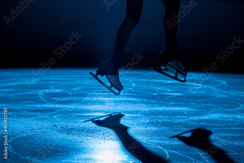 The female athlete in figure skating at the moment of the jump. Women's legs in white skates for figure skating. Practicing skating skills on a dark ice arena with blue light. Close up.
