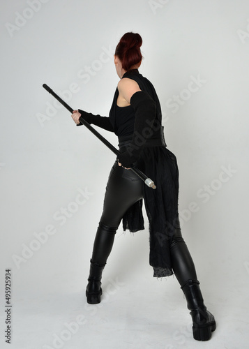 Full length portrait of pretty redhead female model wearing black futuristic scifi leather costume, holding a staff spear weapon. Dynamic standing pose facing away, backwards, white studio background.